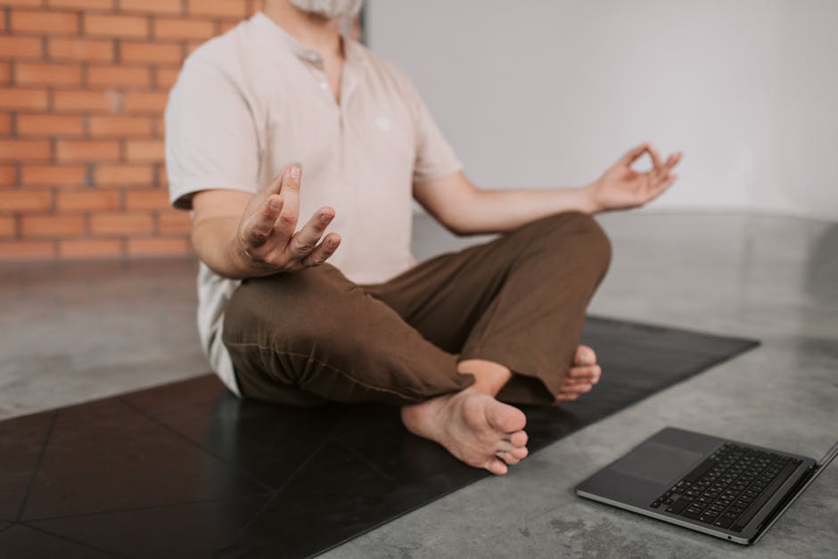 Senior man practicing yoga indoors on a mat, accompanied by a laptop for a virtual session.