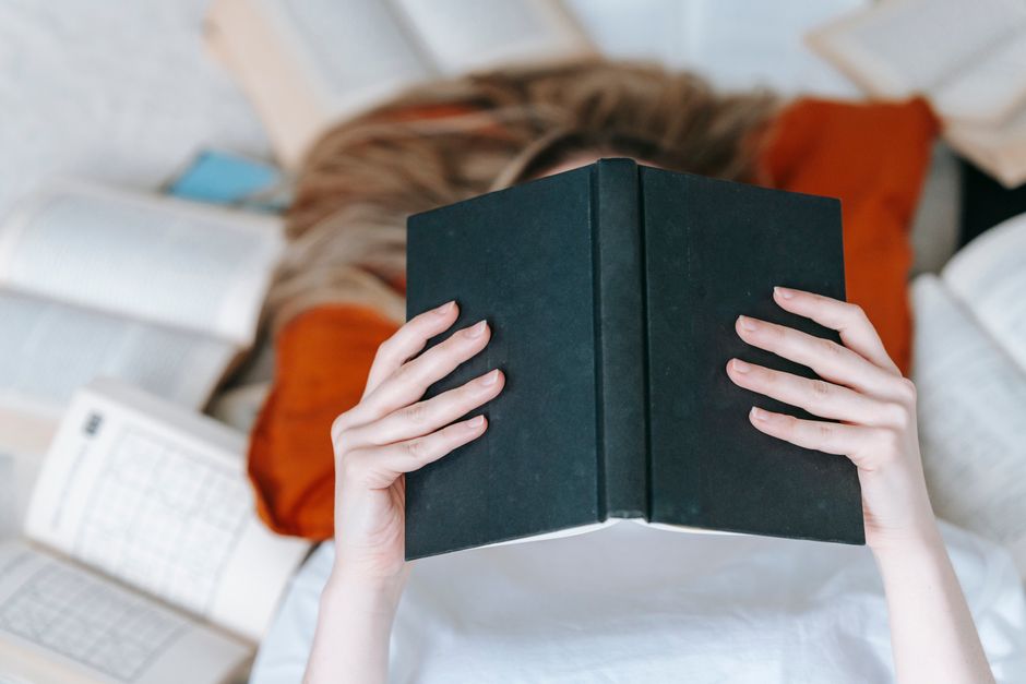 From above of unrecognizable female reading black book with hardcover while lying on floor near various literature in light room