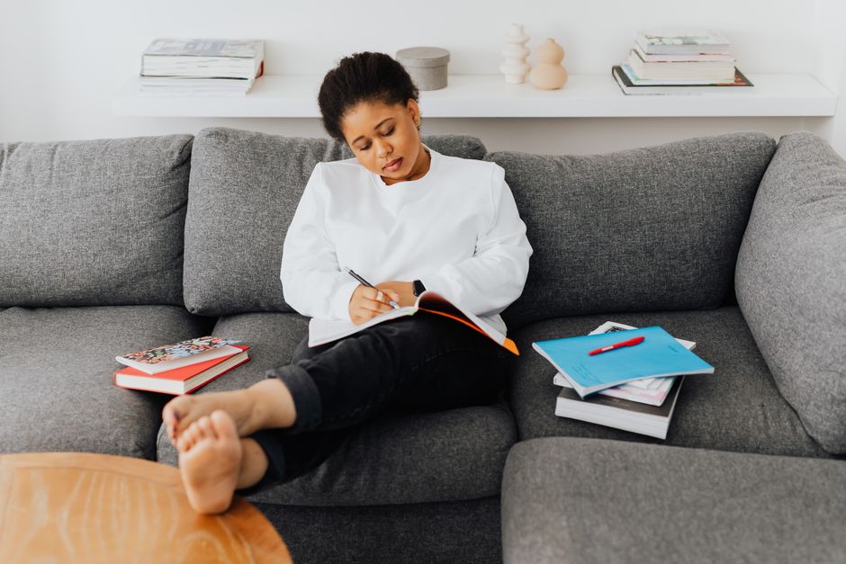 A young woman relaxes on a sofa, writing in a notebook surrounded by books.