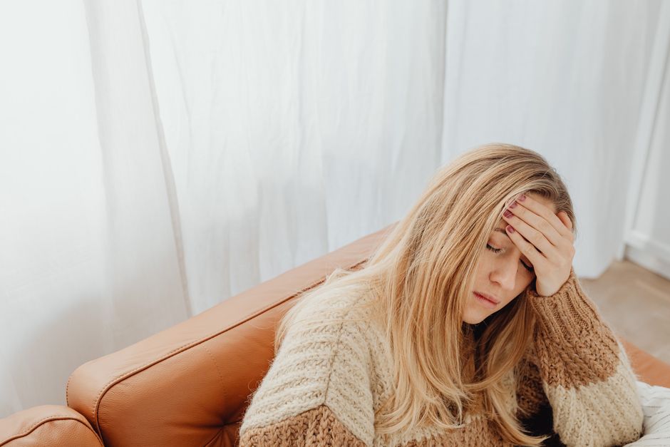 A woman in a knitted sweater sits with a headache on a cozy sofa indoors.